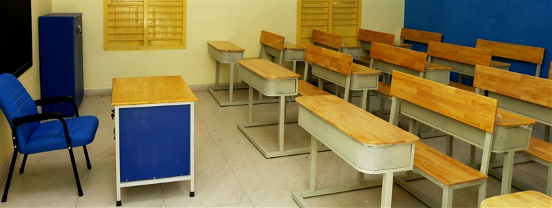 A classroom with a blue chair to the left in front of a wood desk with blue sides and long wooden desks with chairs for the students. Photo by 
Haseeb Modi via Unsplash.